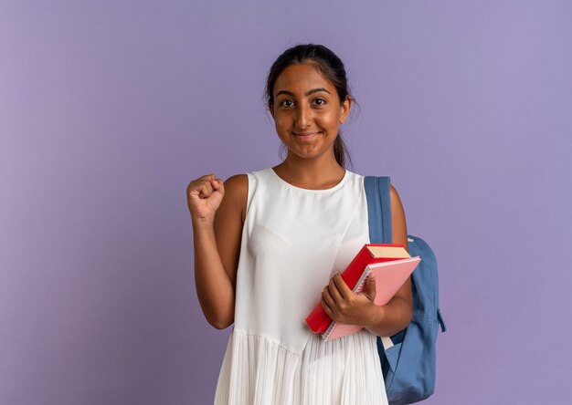 Pleased young schoolgirl wearing back bag holding book and notebook showing yes gesture on purple