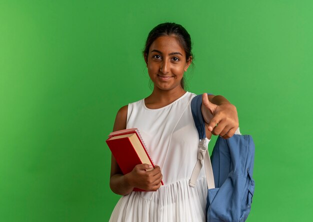 Pleased young schoolgirl wearing back bag holding book with notebook and showing you gesture on green