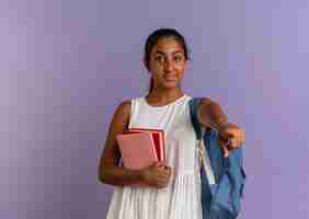 Free photo pleased young schoolgirl wearing back bag holding book with notebook and showing you gesture on purple