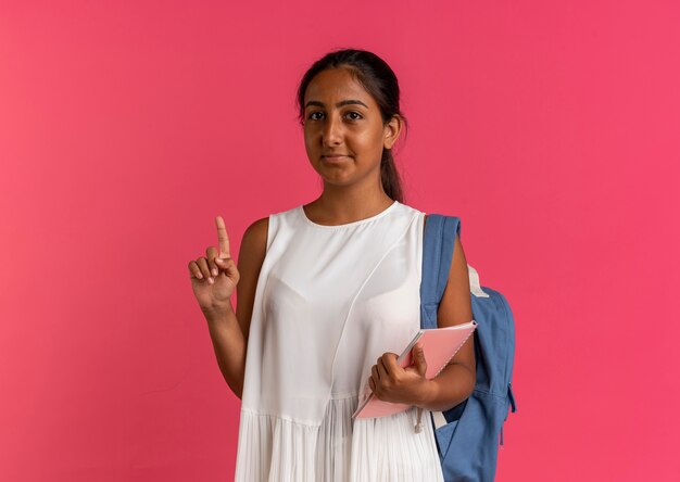 pleased young schoolgirl wearing back bag holding notebook and points at up