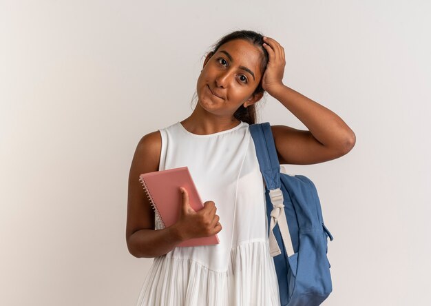 Довольная молодая школьница в задней сумке держит ноутбук и кладет руку на голову на белом