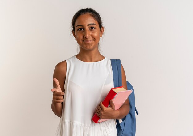 Pleased young schoolgirl wearing back bag holding notebook with book and showing you gesture on white