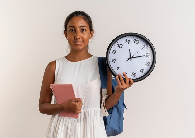 pleased young schoolgirl wearing back bag holding notebook with wall clock