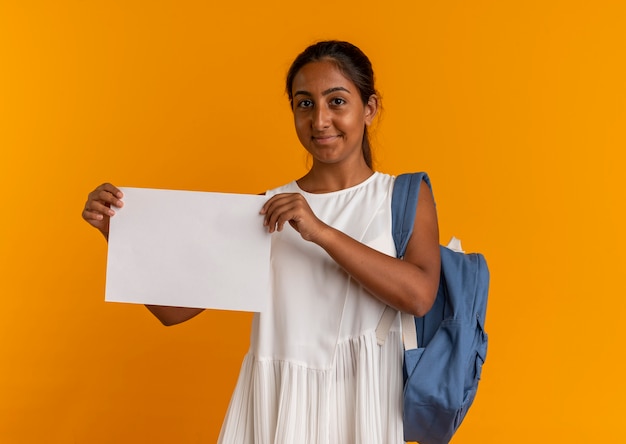 Pleased young schoolgirl wearing back bag holding paper on orange