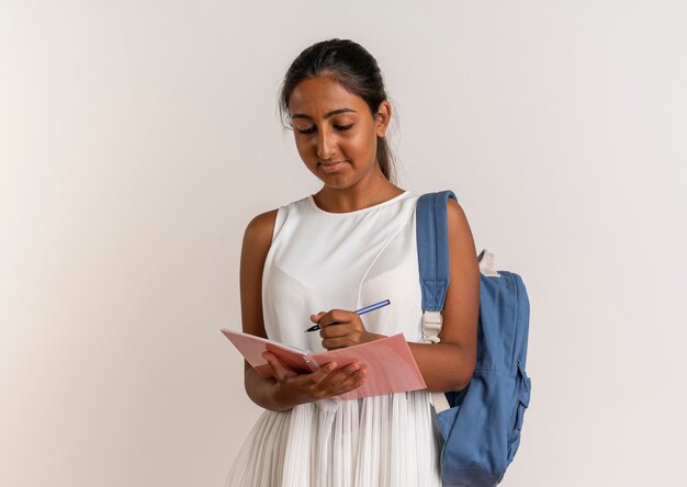 Pleased young schoolgirl wearing back bag holding and write something on notebook on white