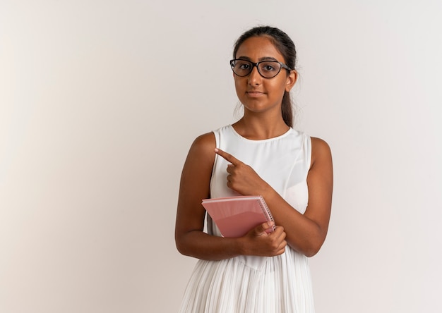 Pleased young schoolgirl wearing glasses holding notebook and points to side isolated on white wall