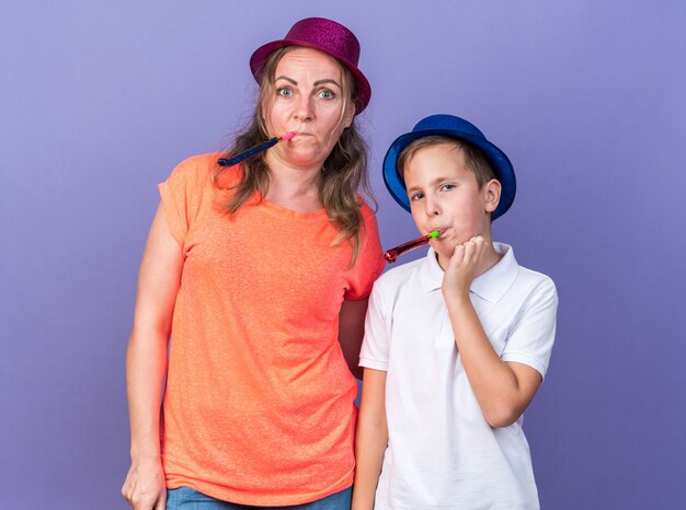 pleased young slavic boy with blue party hat blowing party whistle together with his mother wearing violet party hat isolated on purple wall with copy space