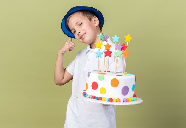 pleased young slavic boy with blue party hat holding birthday cake and gesturing call me sign isolated on olive green wall with copy space