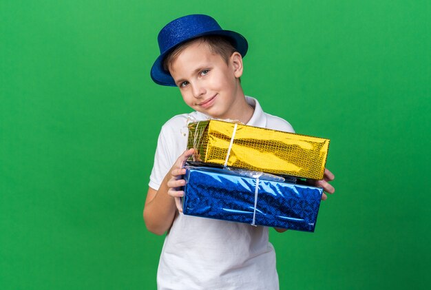 pleased young slavic boy with blue party hat holding gift boxes isolated on green wall with copy space