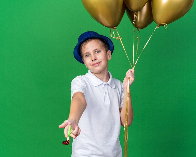 pleased young slavic boy with blue party hat holding helium balloons and party whistle isolated on green wall with copy space