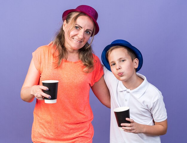 복사 공간이 보라색 벽에 고립 된 보라색 파티 모자를 쓰고 그의 어머니와 함께 서있는 종이 컵을 들고 파란색 파티 모자와 함께 기쁘게 젊은 슬라브 소년