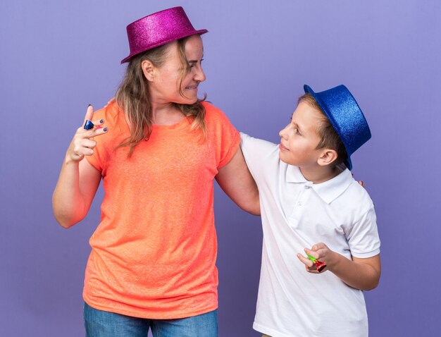 pleased young slavic boy with blue party hat holding party whistles together with his mother wearing violet party hat isolated on purple wall with copy space