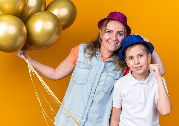 pleased young slavic boy with blue party hat keeping fist up and standing with his mother wearing purple party hat holding helium balloons isolated on orange wall with copy space