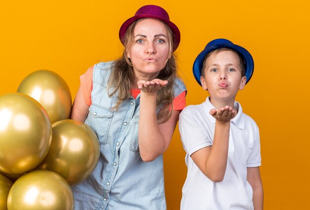 pleased young slavic boy with blue party hat sending kiss with hand and standing with his mother wearing purple party hat holding helium balloons isolated on orange wall with copy space