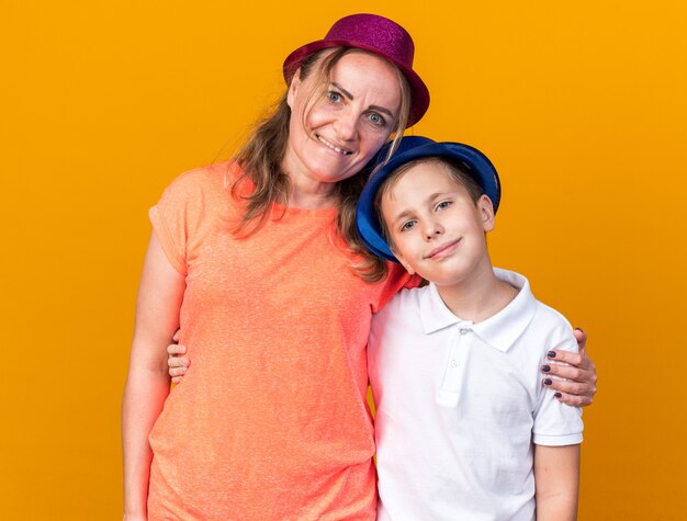 pleased young slavic boy with blue party hat standing with his mother wearing purple party hat isolated on orange wall with copy space