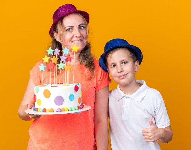 довольный молодой славянский мальчик в синей праздничной шляпе, стоящий с его матерью в фиолетовой праздничной шляпе и держащий именинный торт, изолированный на оранжевой стене с копией пространства