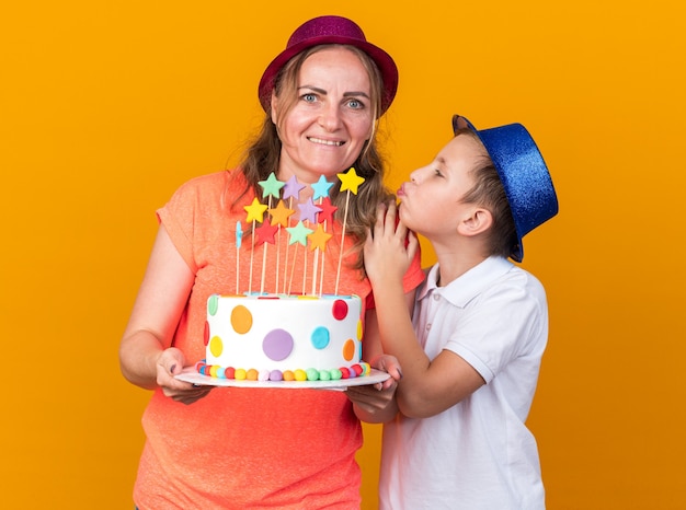 pleased young slavic boy with blue party hat trying to kiss his mother wearing purple party hat and holding birthday cake isolated on orange wall with copy space