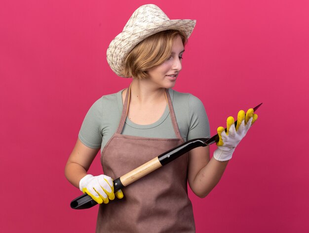 Довольная молодая славянская женщина-садовник в садовой шляпе и перчатках держит и смотрит на лопату на розовом