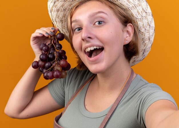 Pleased young slavic female gardener wearing gardening hat holding grapes looking at camera taking selfie