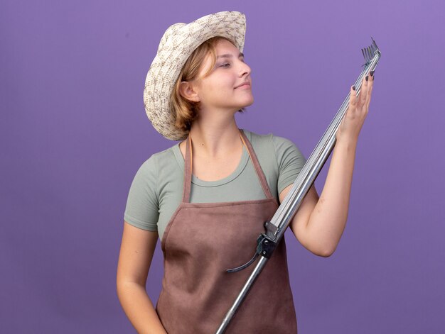 Pleased young slavic female gardener wearing gardening hat holding and looking at leaf rake on purple