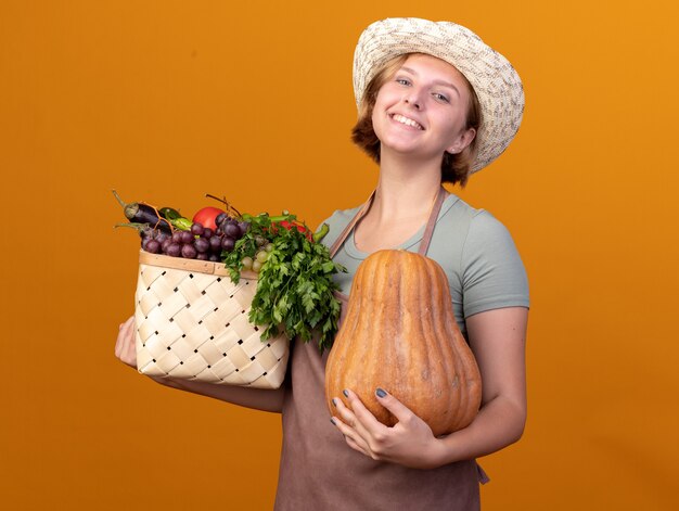 Pleased young slavic female gardener wearing gardening hat holding vegetable basket and pumpkin on orange