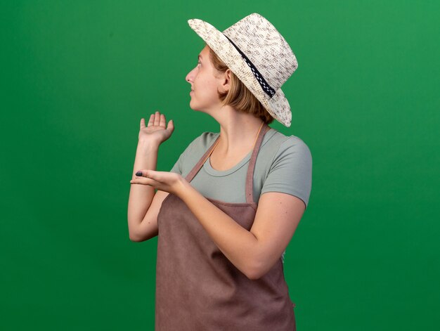 Pleased young slavic female gardener wearing gardening hat looking and pointing back on green