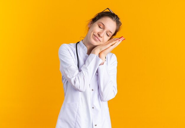 의사복을 입은 젊은 슬라브계 소녀가 청진기를 들고 주황색 벽에 복사공간이 있는 손에 머리를 얹고 있다