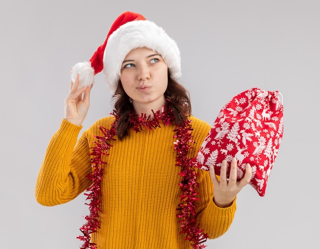 산타 모자와 크리스마스 선물 가방을 들고 목에 갈 랜드와 복사 공간 흰색 배경에 고립 된 측면을보고 기쁘게 젊은 슬라브 소녀