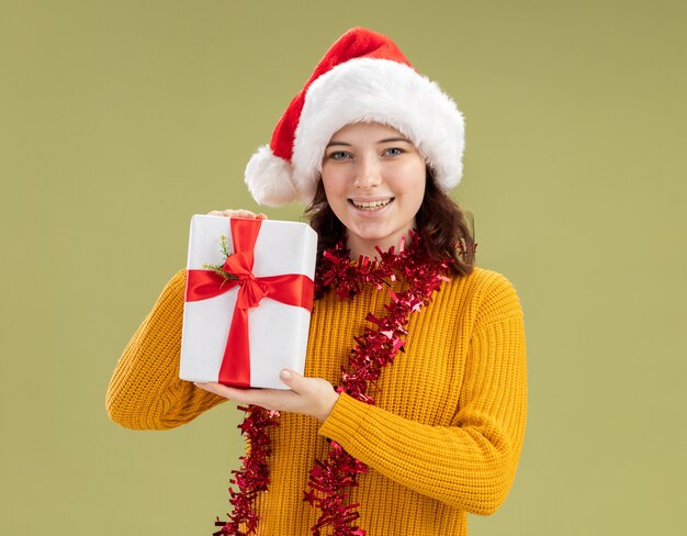 Pleased young slavic girl with santa hat and with garland around neck holding christmas gift box isolated on olive green wall with copy space