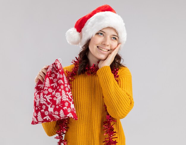 Pleased young slavic girl with santa hat and with garland around neck puts hand on face and holds christmas gift bag looking at side isolated on white wall with copy space