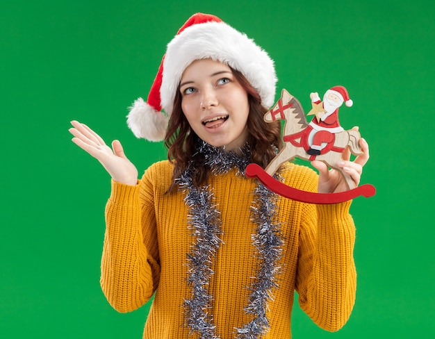 산타 모자와 목에 갈 랜드가있는 기쁘게 젊은 슬라브 소녀가 혀를 내밀고 흔들 목마 장식에 산타를 보유하고 있습니다.
