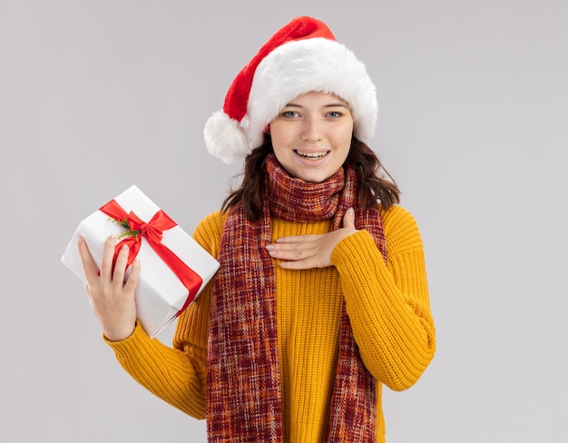 pleased young slavic girl with santa hat and with scarf around neck puts hand on chest and holds christmas gift box isolated on white background with copy space