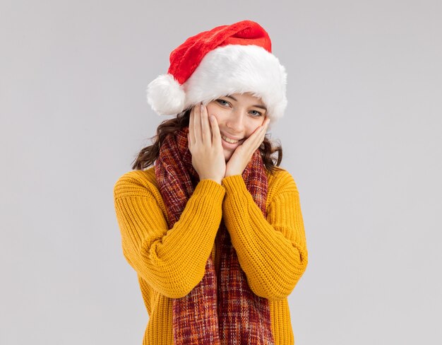 Pleased young slavic girl with santa hat and with scarf around neck puts hands on face 