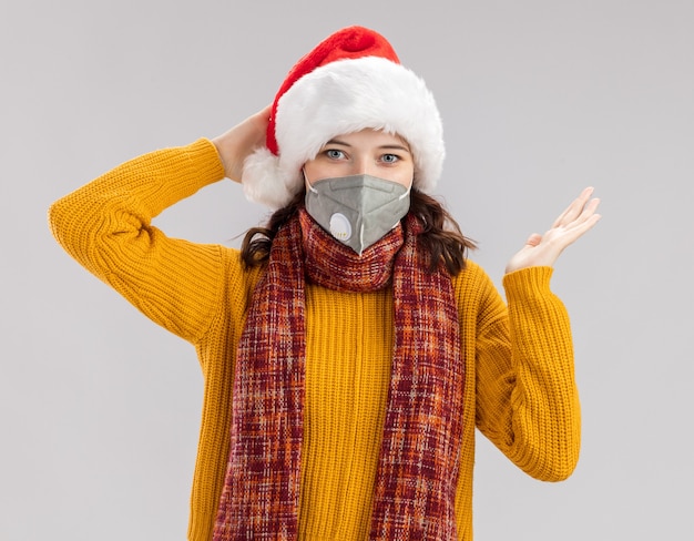 pleased young slavic girl with santa hat and with scarf around neck wearing medical mask keeps hand open isolated on white background with copy space