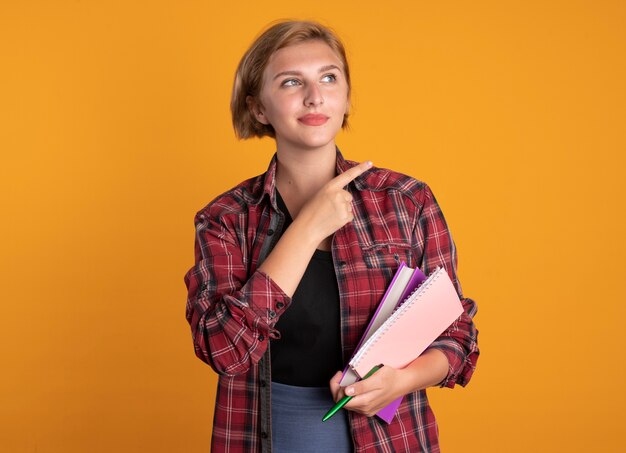 Pleased young slavic student girl holds pen book and notebook looking and pointing at side 