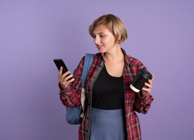 Довольная молодая славянская студентка в рюкзаке держит бумажный стаканчик и смотрит на телефон