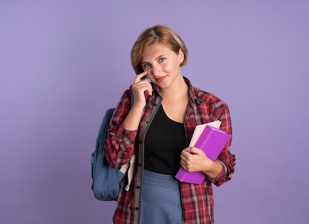 Pleased young slavic student girl wearing backpack puts finger on eyelid holds book and notebook 