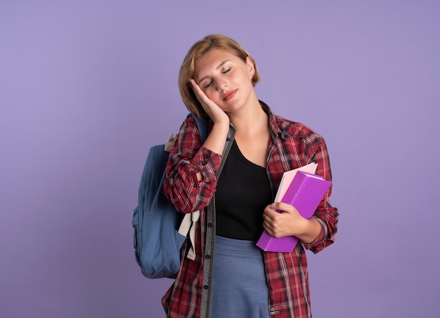 Pleased young slavic student girl wearing backpack puts hand on face holds book and notebook 