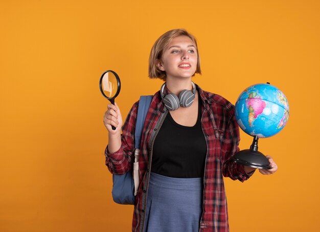 Pleased young slavic student girl with headphones wearing backpack holds globe and magnifying glass 