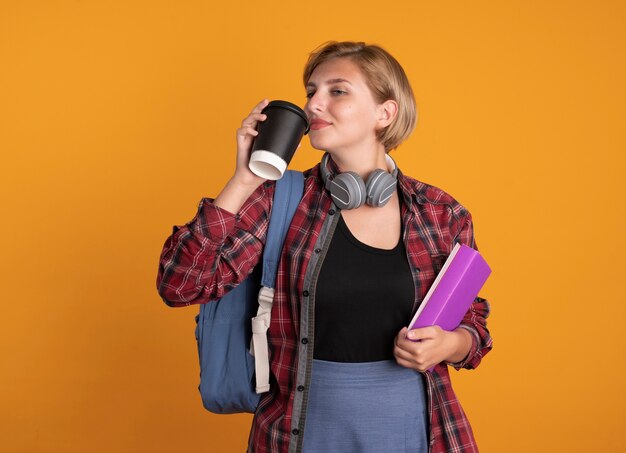 배낭을 착용하는 헤드폰으로 기쁘게 젊은 슬라브 학생 소녀는 종이 컵 책과 노트북을 보유하고 있습니다.