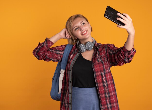 Pleased young slavic student girl with headphones wearing backpack puts hand on head holding and looking at phone 