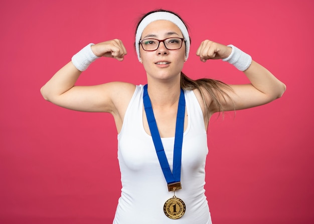 Pleased young sporty girl in optical glasses with gold medal around neck wearing headband and wristbands tenses biceps Free Photo