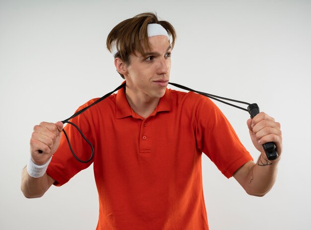 Pleased young sporty guy looking at side wearing headband with wristband holding jump rope on neck
