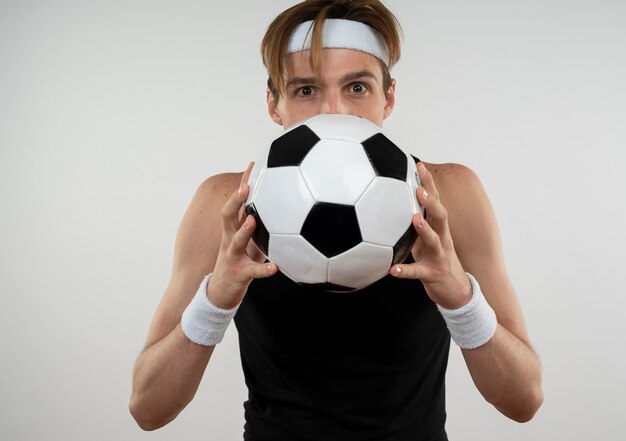 Pleased young sporty guy wearing headband and wristband covered face with ball isolated on white