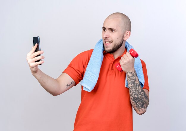 Pleased young sporty man holding dumbbell with towel on shoulder take a selfie isolated on white wall