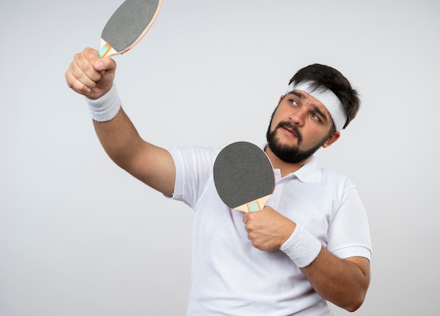 Foto gratuita soddisfatto giovane sportivo che guarda al lato che indossa la fascia e il braccialetto che tiene la racchetta da ping pong isolata sul muro bianco