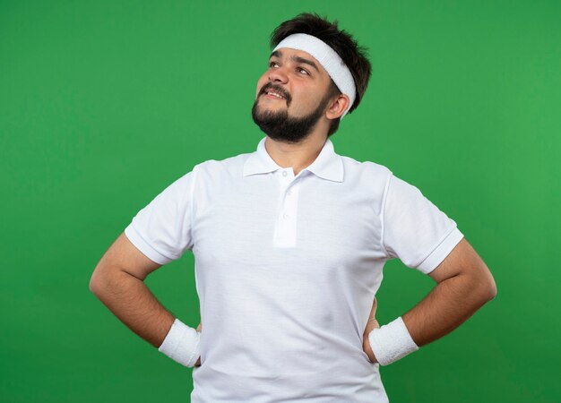 Pleased young sporty man looking at side wearing headband and wristband putting hands on hip isolated on green