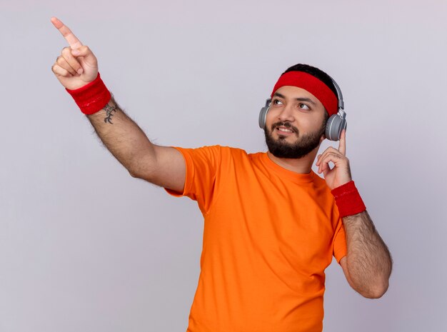 Pleased young sporty man looking at side wearing headband and wristband with headphones points