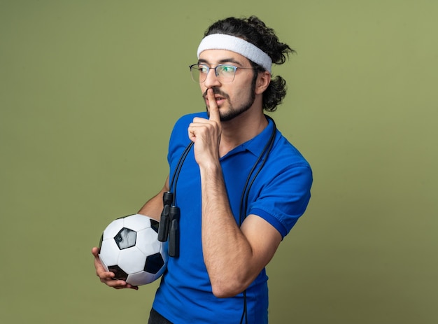 Довольный молодой спортивный мужчина с повязкой на голову с браслетом и скакалкой на плече держит мяч, показывая жест тишины
