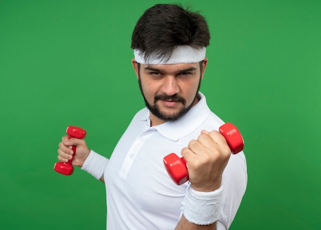Pleased young sporty man wearing headband and wristband exercising with dumbbells isolated on green wall